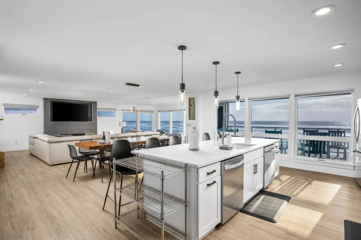 This image captures an expansive, sunlit open-plan living space where the kitchen island, dining area, and comfortable lounge seating all flow together. Huge windows offer unobstructed ocean views, and the space is filled with natural light, making it perfect for gatherings and shared meals.