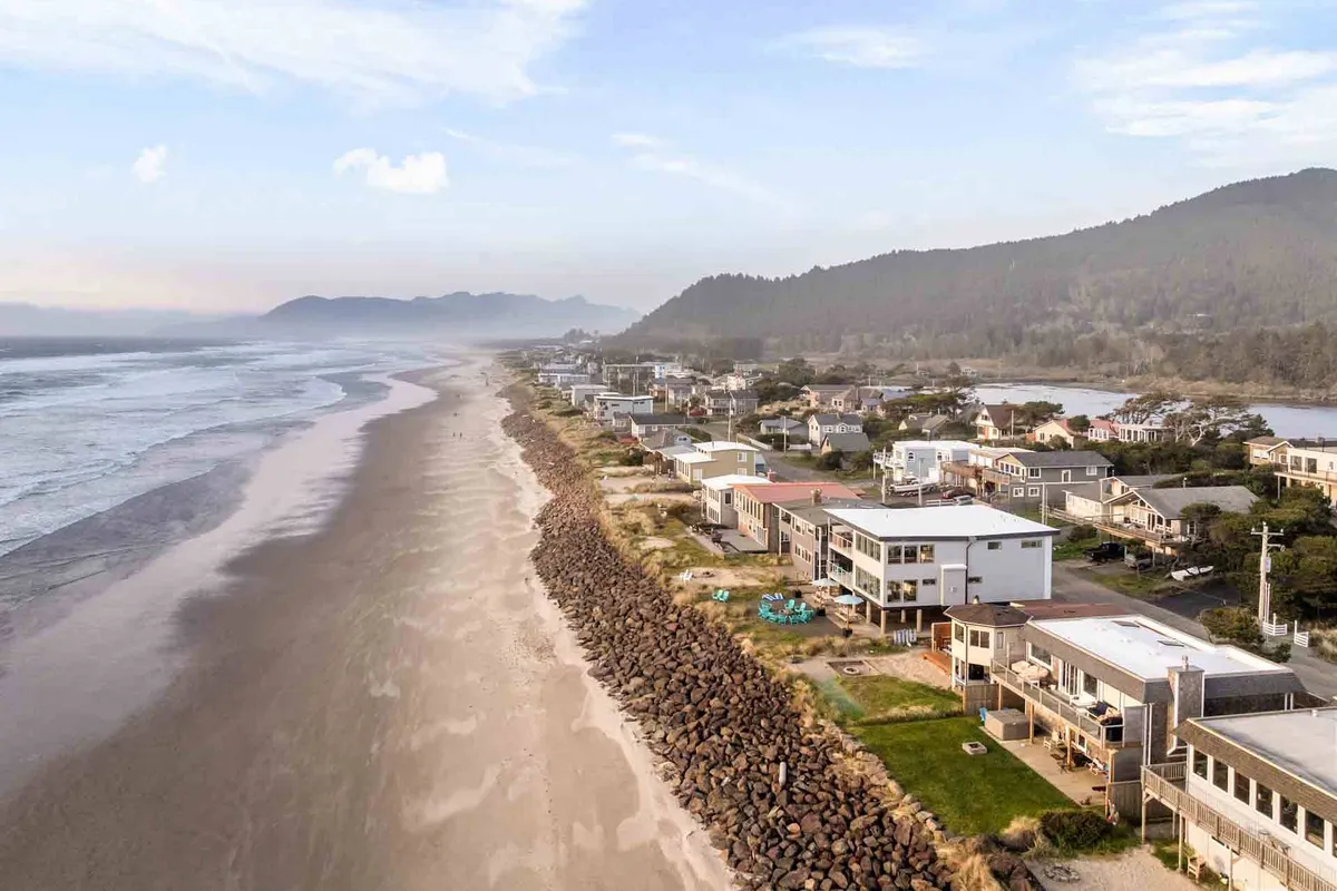 A scenic stretch of Rockaway Beach is captured with beachfront houses lining the edge, a broad expanse of sandy shoreline, rocky outcroppings, and gentle waves rolling in. The view is sweeping, highlighting the inviting and natural coastline perfect for dog walks and seaside adventure.