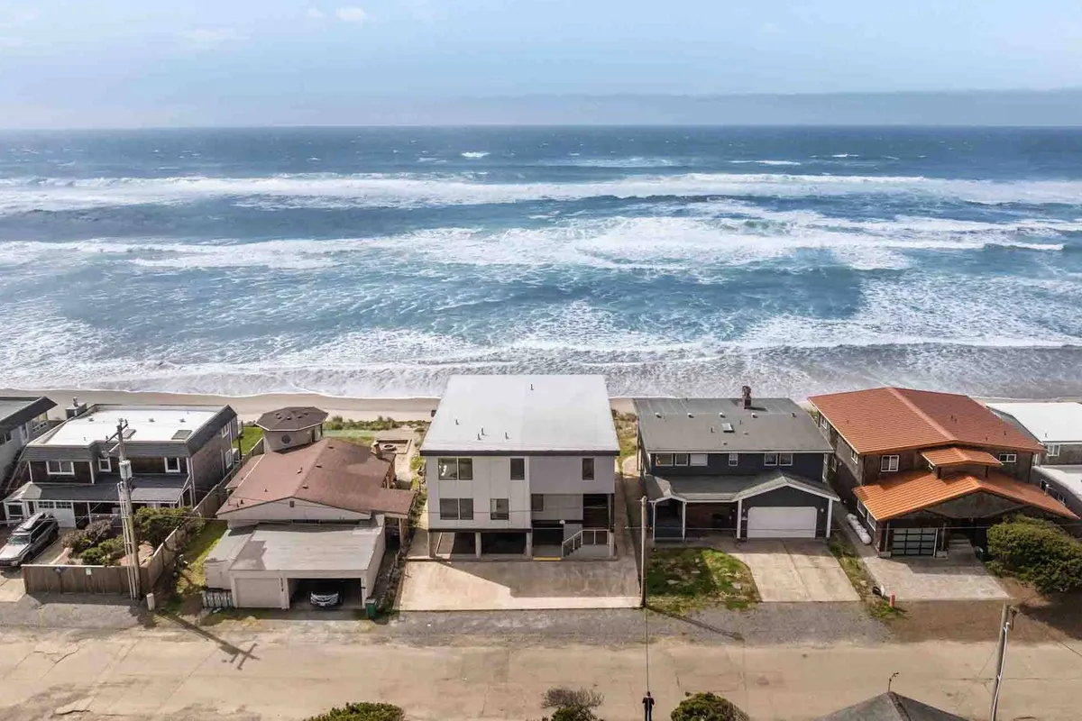 This image shows a stretch of sandy Rockaway Beach with beachfront houses lining the shore. The ocean waves roll gently onto the sand, offering a clear sense of the direct beach access available to guests. Families and friends can be imagined strolling, playing, or relaxing just steps from their accommodations.