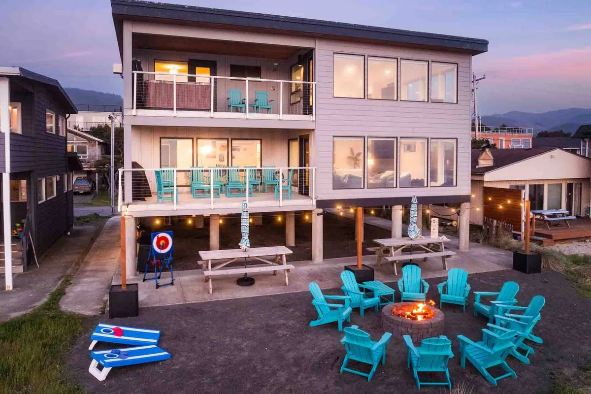 The image showcases a stunning two-story beach house with expansive outdoor spaces, including a cozy fire pit surrounded by colorful chairs, picnic tables, and inviting seating areas—all just steps from the Oregon Coast. The sunset light creates a warm, communal atmosphere perfect for gathering with friends and family.