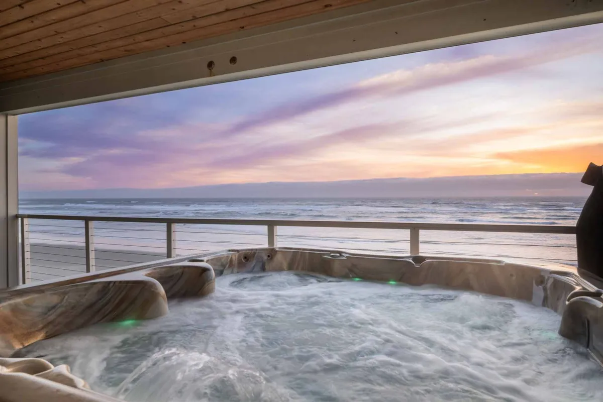 An exterior shot focusing on a bubbling hot tub set against the backdrop of the ocean and a glowing sunset sky. The inviting waters of the hot tub are complemented by comfortable seating and the serene natural panorama, promising ultimate leisure and breathtaking vistas.