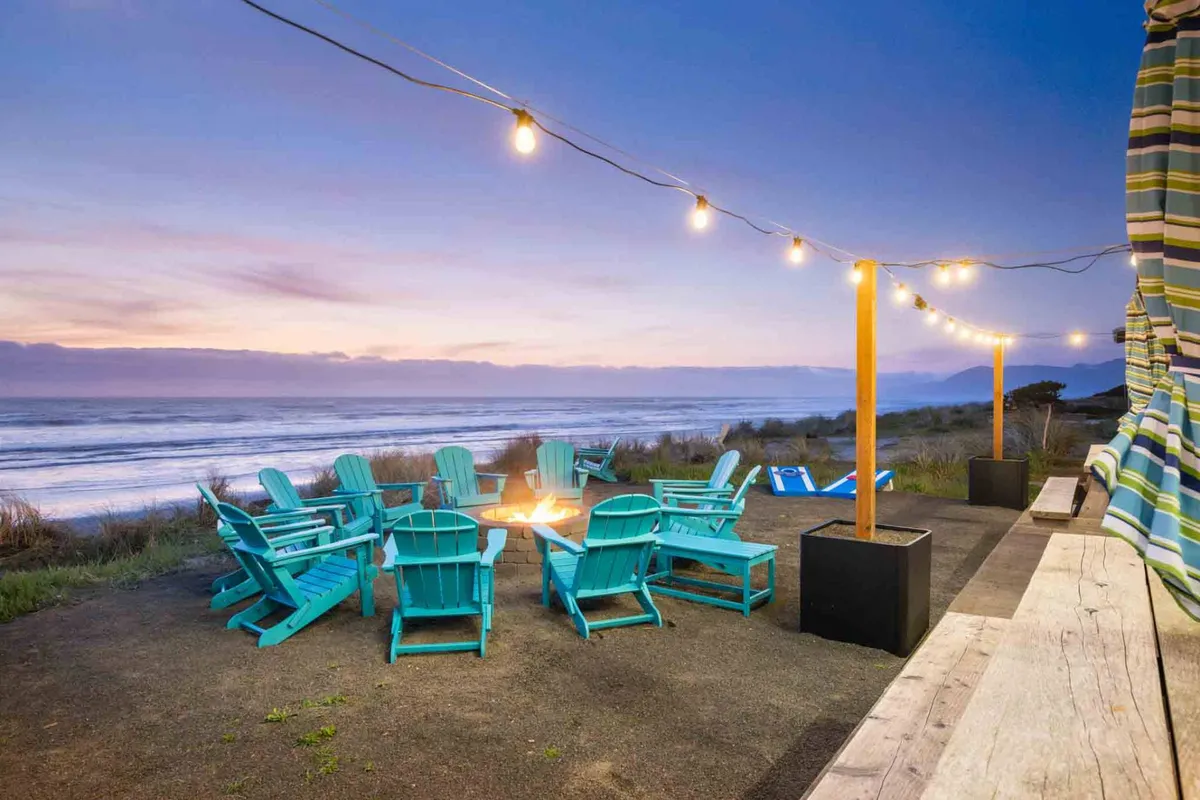 An inviting beachfront fire pit surrounded by Adirondack chairs, string lights overhead, and a stunning ocean view in the background. The sandy area and relaxed setup encourage gathering and socializing right by the beach.