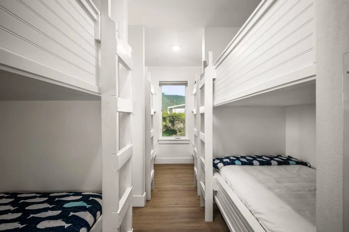 A cheerful bunk room with sturdy bunk beds dressed in textured bedding, a window with a view, and a playful, communal design. The space is inviting for children or groups to bond, play, and rest together.