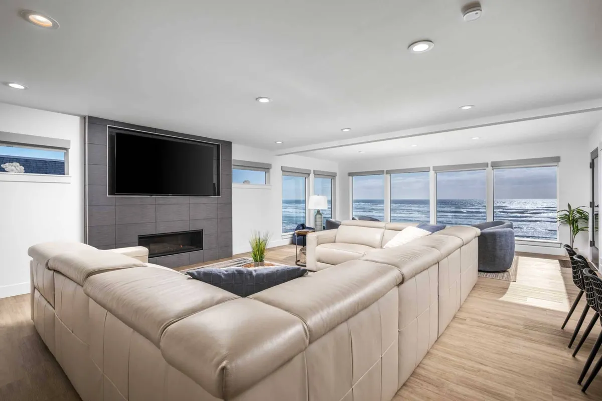 A spacious interior with a large sectional sofa positioned to take advantage of panoramic ocean views through wide windows. Indoors, there are cozy elements like indoor plants and a TV, but the main feature is the visual access to the stunning coastline just outside.