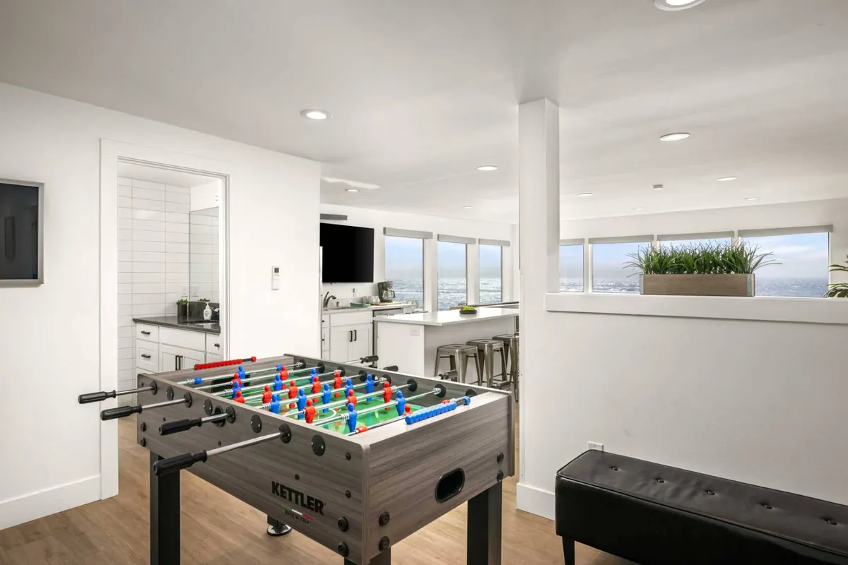 The image captures the entertainment loft featuring a foosball table in the foreground, with a kitchen area and expansive windows offering striking ocean views. The scene buzzes with a sense of fun and energy, inviting socializing and playful competition in a setting that feels both vibrant and relaxed.