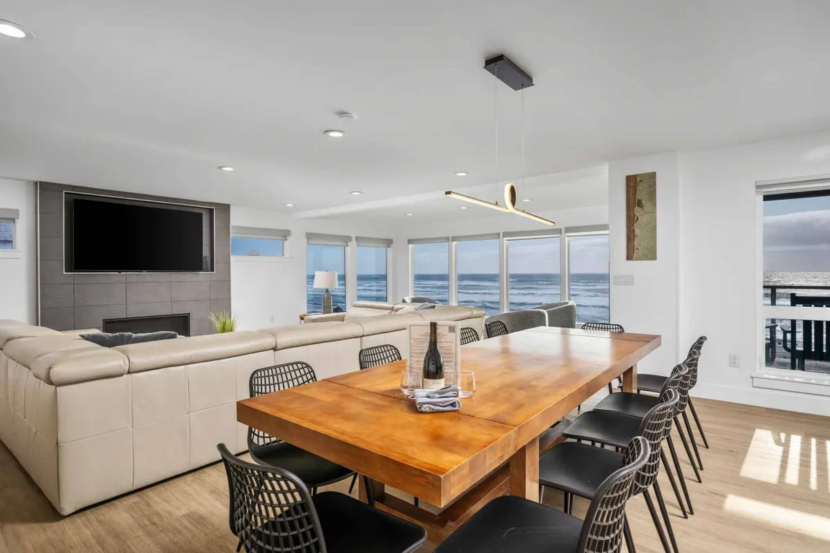 A generous dining table is set beside wide windows that overlook the ocean, surrounded by a plush sectional sofa and modern lighting. The space is designed for both intimate family dinners and festive social gatherings.