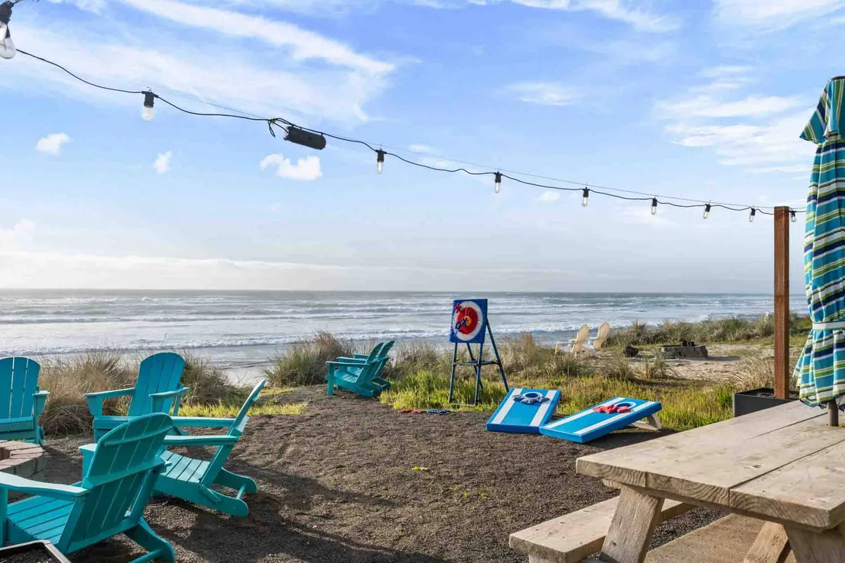 A casual beachfront setting with Adirondack chairs, an outdoor table, string lights, ocean view, and a bean bag toss game set up on the sand. The area is lively and ready for outdoor games and social fun right at the edge of the beach.