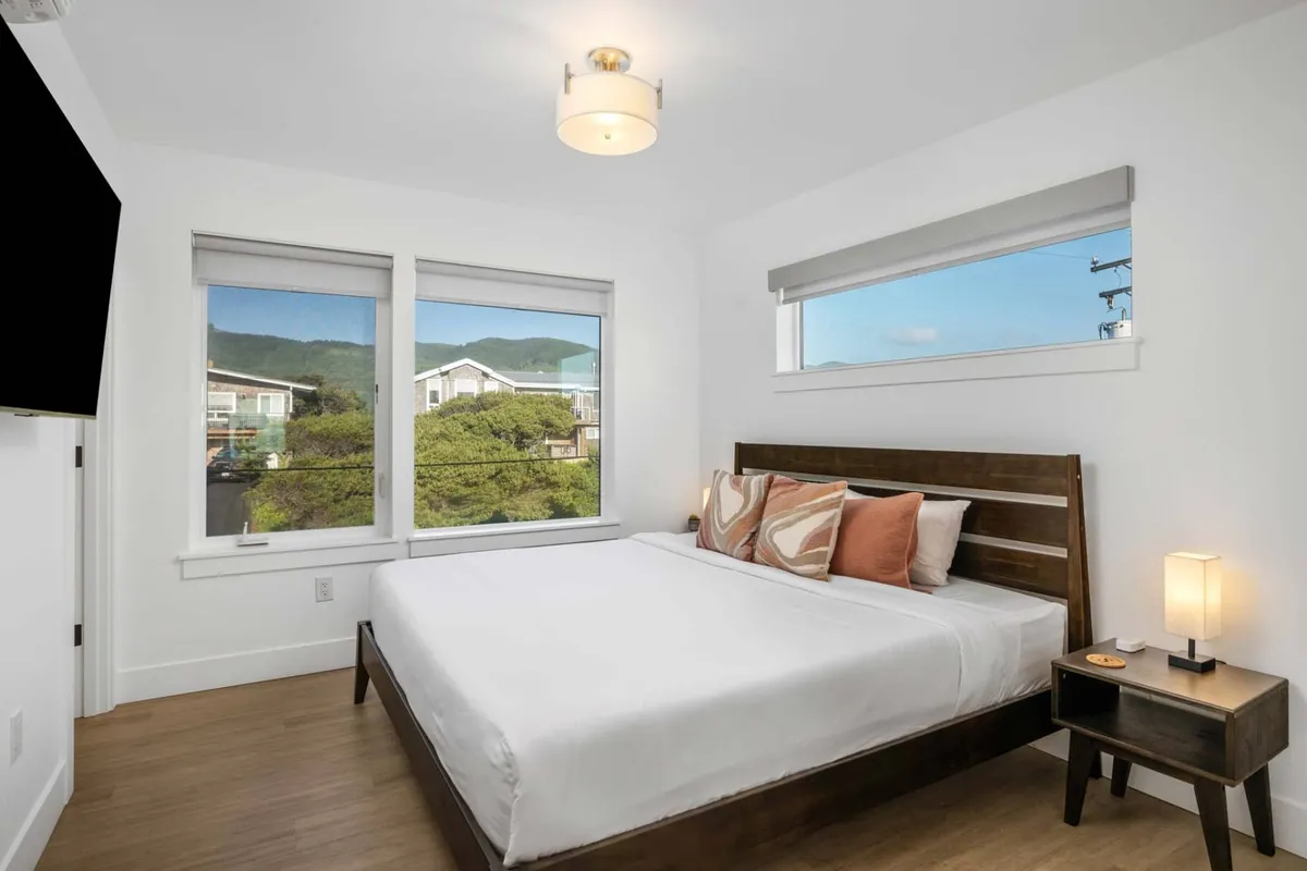 A comfortable bedroom with a stylish bed adorned with decorative pillows, complemented by large windows that invite natural light and offer a scenic view. A nightstand with a lamp enhances the cozy, welcoming feel of the room.